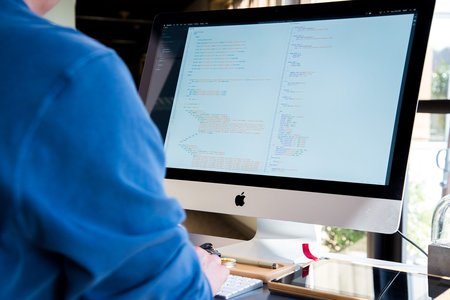 Person in blauer Kleidung arbeitet an einem Apple-Monitor mit geöffnetem Code-Editor